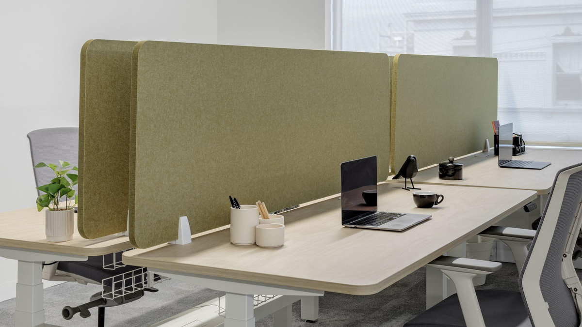A modern office setup with light wood desks, gray ergonomic chairs, and tall beige fabric partition walls. A laptop, small plant, and desk accessories are arranged on the desk in a bright, clean workspace.