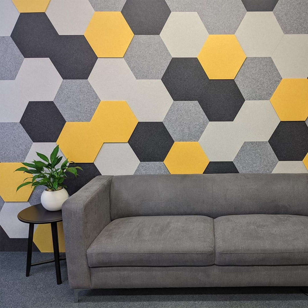 Honeycomb acoustic tiles in grey and yellow on a wall with a modern grey sofa and potted plant in front.