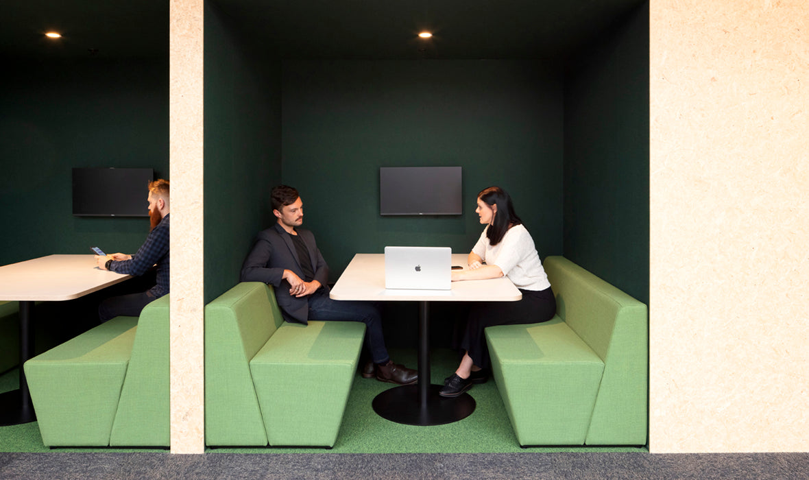 Two professionals engaged in discussion at a modern office booth while another person uses a phone in the background.